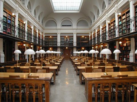 University library reading room with wooden desks, white reading lamps and two floors of bookshelves