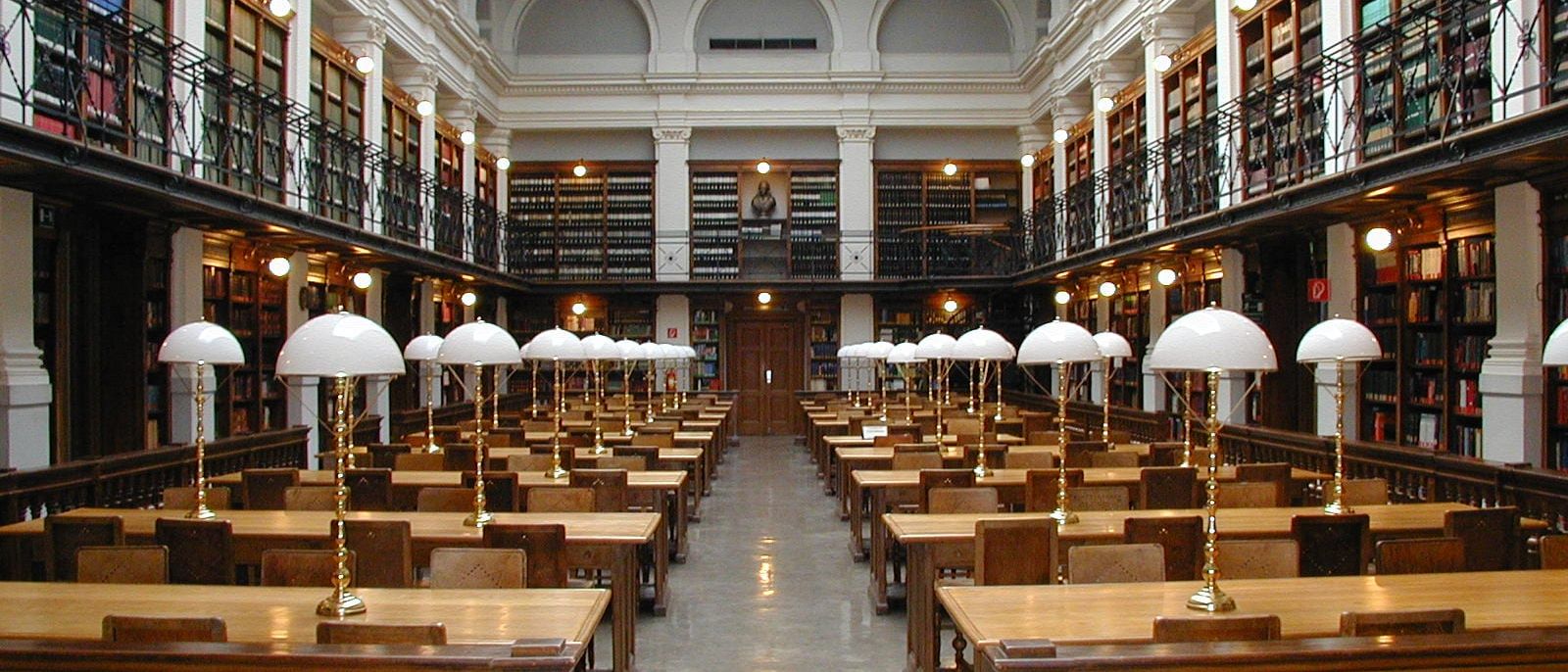 University library reading room with wooden desks, white reading lamps and two floors of bookshelves